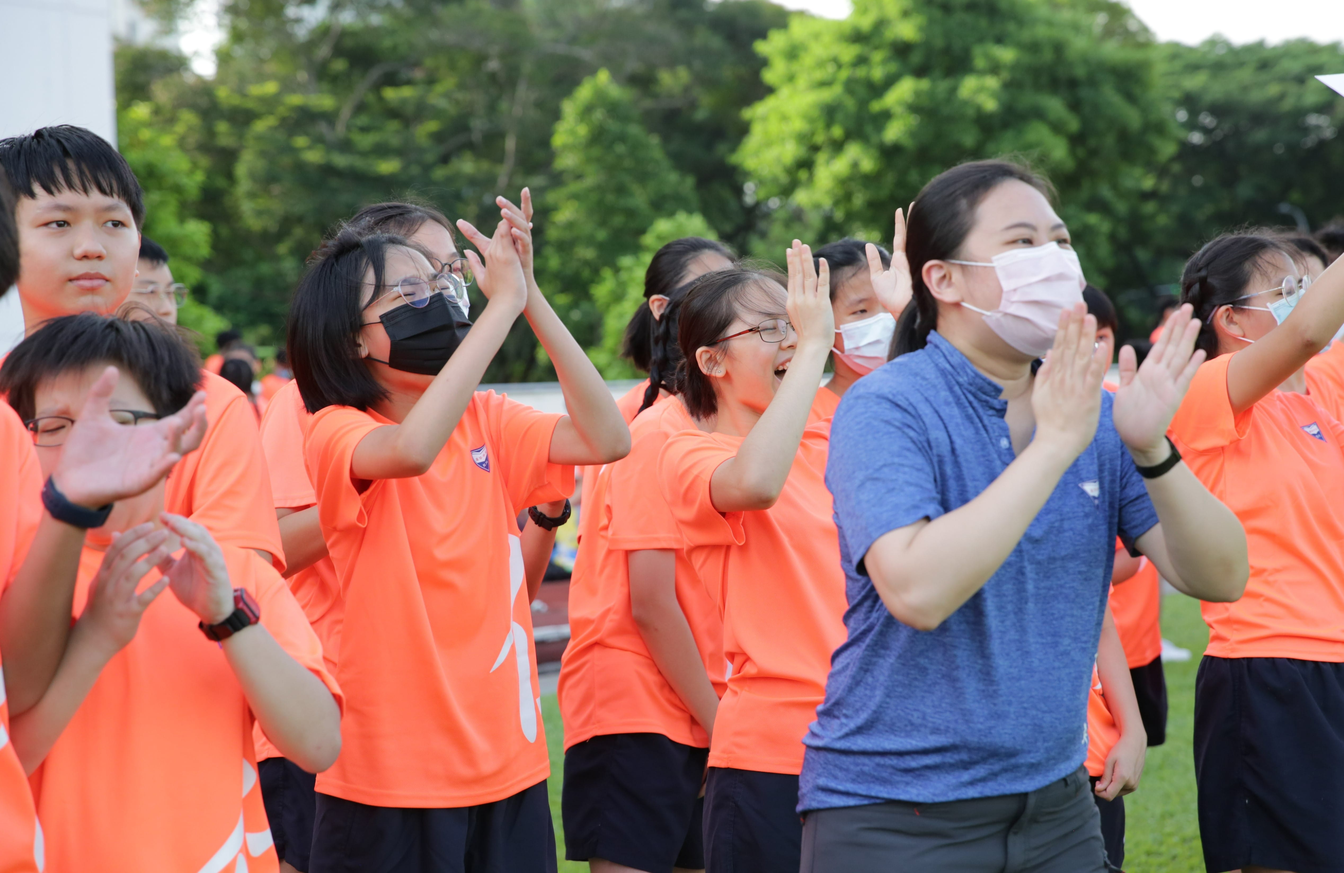A group of students in orange shirts and an adult in blue, all wearing masks, clap and move outdoors near trees.